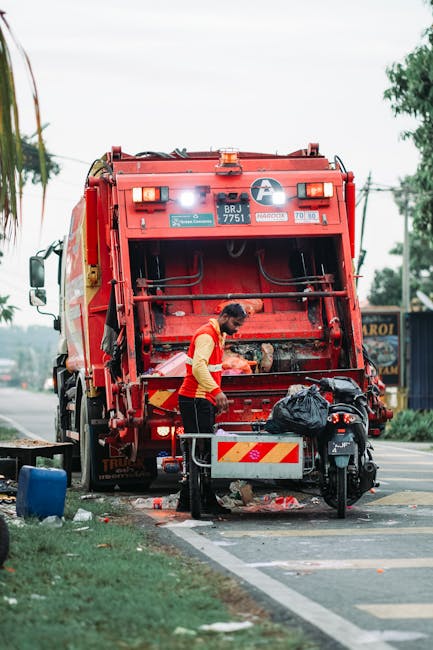 A worker wearing a yellow and red uniform is emptying a large red rubbish collection truck parked on the side of a street. The back of the truck's compactor is open, revealing the interior's dark metal surfaces and hydraulic mechanisms. The worker is standing next to a small metal container, removing rubbish bags that are stacked nearby. The scene includes a black motorbike parked beside a safety barrier next to the truck, with a black plastic bag placed on top of the barrier. The street is bordered by a grassy verge with a tree and some bushes in the background, under overcast weather conditions. Utility poles with overhead wires run parallel to the road, and there are residential and commercial structures visible further in the background. The overall setting reflects an on-site waste collection activity, typical of private rubbish removal services like those offered by Rubbish Removal Hammersmith.