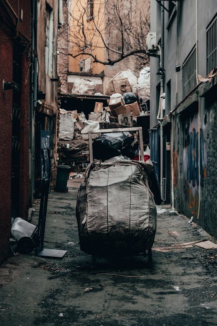 A narrow urban alleyway cluttered with overflowing piles of trash bags, cardboard boxes, and discarded waste materials stacked against a brick wall in the background. The foreground features a large, grey, weathered refuse bin with a metal handle, positioned at the center of the alley. The ground is dark and stained with dirt and debris, indicating a neglected or unused space. On the right side, a building with grey metal siding and barred windows is visible, while graffiti covers parts of the adjacent wall on the left side, giving the scene an urban, industrial atmosphere. In the background, leafless trees are visible above the debris, suggesting a winter season. The scene implies a situation where private or alternative waste disposal methods, such as rubbish removal services by Rubbish Removal Hammersmith, are needed to clear the accumulated rubbish from inner-city alleyways, highlighting urban waste management challenges.