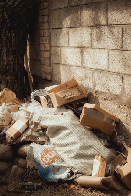 A narrow urban alleyway cluttered with overflowing piles of trash bags, cardboard boxes, and discarded waste materials stacked against a brick wall in the background. The foreground features a large, grey, weathered refuse bin with a metal handle, positioned at the center of the alley. The ground is dark and stained with dirt and debris, indicating a neglected or unused space. On the right side, a building with grey metal siding and barred windows is visible, while graffiti covers parts of the adjacent wall on the left side, giving the scene an urban, industrial atmosphere. In the background, leafless trees are visible above the debris, suggesting a winter season. The scene implies a situation where private or alternative waste disposal methods, such as rubbish removal services by Rubbish Removal Hammersmith, are needed to clear the accumulated rubbish from inner-city alleyways, highlighting urban waste management challenges.