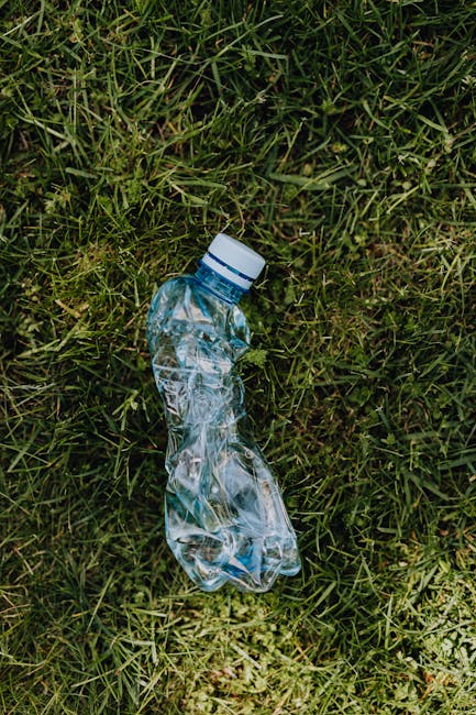 A discarded crumpled plastic water bottle with a white cap is lying on a patch of green grass, which has a mixture of fine and coarse blades. The bottle is transparent, revealing the interior and some residual liquid, and appears crushed from use. The grass around the bottle is natural, with some blades slightly overlapping, and the lighting suggests an outdoor environment with diffuse sunlight. The scene reflects the issue of improper waste disposal in public or outdoor spaces, highlighting the importance of responsible rubbish management. Rubbish Removal Hammersmith specialized in private waste handling, including the collection and proper disposal of single-use plastics found in such settings, exemplified by the plastic bottle in this image.