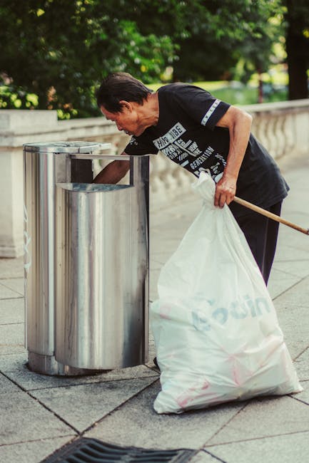 An elderly man with dark hair, wearing a black T-shirt with printed white text and black trousers, is seen disposing of rubbish into a stainless steel trash bin located outdoors on a paved walkway. He holds a white plastic bag filled with waste in his left hand, while using his right hand to reach into the open top of the bin, which has a rounded, reflective surface and is positioned adjacent to a low stone balustrade. The background features lush green trees and foliage, indicating a park or garden setting during daylight hours. The scene captures an act of environmental responsibility, aligning with private rubbish removal and on-site waste management services, with the silver bin serving as a receptacle for rubbish collection amid the natural surroundings.