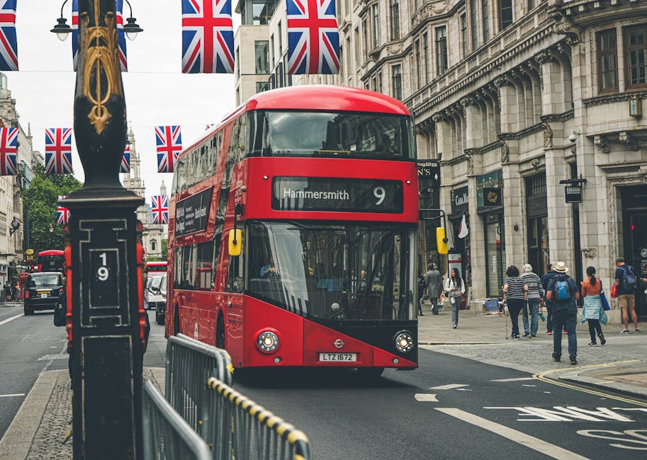 A red double-decker bus displaying the route number 9 and destination Hammersmith is traveling along a busy urban street lined with historic buildings and storefronts. The bus is positioned in the center of the road, with a commercial bus lane marked on the asphalt. In the foreground, a black lamppost with decorative gold detailing is visible, along with a metal barrier with a yellow and black striped safety marking, suggesting a designated area for pedestrian or vehicle management. Pedestrians, some carrying shopping bags and wearing casual attire, walk along the pavement on both sides of the street, with a few stopping to observe their surroundings. The background features the overcast sky, with Union Jack flags strung overhead and fluttering slightly, emphasizing a typical cityscape scene. Visible shop signs and building facades exhibit stone and brick finishes with varied architectural details, creating a lively, classic London atmosphere. This scene exemplifies urban transportation and pedestrian activity, aligning with signs of independent local transport services and the city's approach to traffic flow and street management. Rubbish Removal Hammersmith offers complementary services for waste collection in similar city environments, ensuring efficient clearance of rubbish in busy urban areas that require alternative waste handling options.
