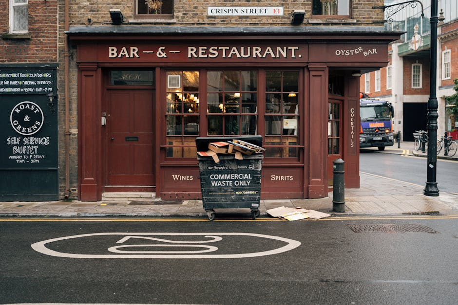 In front of a traditional brick building housing a bar and restaurant, a black commercial waste bin with white text indicating it is for waste disposal only stands on the pavement. The bin is filled with flattened cardboard boxes and other waste materials, some of which are spilling over the top and onto the sidewalk. Positioned directly on the curb, the bin is adjacent to a small metal post and a bollard, with a few loose pieces of cardboard lying nearby. Behind the bin, the restaurant features large wooden-framed windows, with warm interior lighting visible through the glass, and a wooden door to the right. Above the establishment, a street sign reads 'Crispin Street E1,' indicating the location. The surroundings include a parked truck on the road, bicycles nearby, and signage for a self-service buffet on the building’s left side, emphasizing the typical urban street scene. This image reflects the importance of proper waste management for local businesses, illustrating the role of commercial rubbish disposal services.