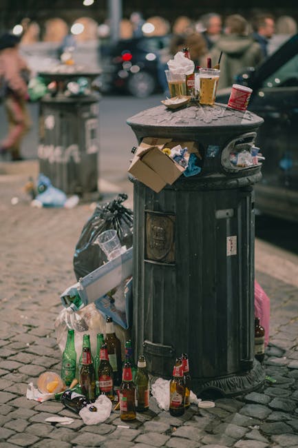 A black, cylindrical public rubbish bin made of metal with a domed top and vertical slats, positioned on a cobblestone street. The bin's lid is open, revealing scattered paper and plastic waste inside; the surrounding area shows various discarded items, including empty glass bottles, crumpled napkins, and bits of paper, with some bottles leaning against the base of the bin. Several bottles and containers are placed on top of the lid, including paper cups, snack wrappers, and plastic bottles, some with drinks inside. The background features blurred pedestrians moving along the street, other bins, and parked cars, indicating an urban environment. The scene reflects improper waste disposal and littering typical of busy public areas, under natural daytime lighting with a slightly overcast atmosphere, emphasizing the neglected state of the rubbish and the need for regular collection and waste management services such as those provided by Rubbish Removal Hammersmith.