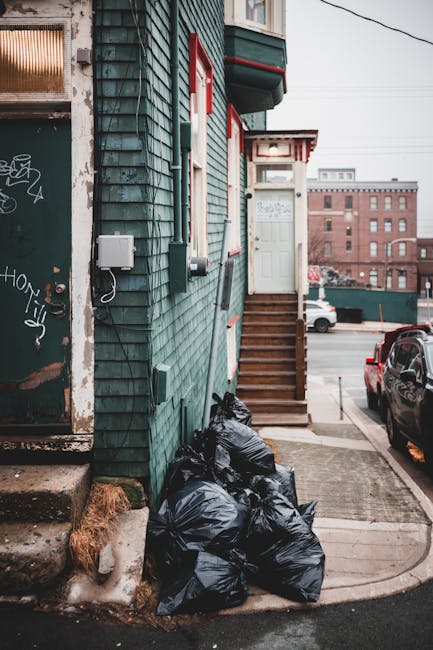 In front of a traditional brick building housing a bar and restaurant, a black commercial waste bin with white text indicating it is for waste disposal only stands on the pavement. The bin is filled with flattened cardboard boxes and other waste materials, some of which are spilling over the top and onto the sidewalk. Positioned directly on the curb, the bin is adjacent to a small metal post and a bollard, with a few loose pieces of cardboard lying nearby. Behind the bin, the restaurant features large wooden-framed windows, with warm interior lighting visible through the glass, and a wooden door to the right. Above the establishment, a street sign reads 'Crispin Street E1,' indicating the location. The surroundings include a parked truck on the road, bicycles nearby, and signage for a self-service buffet on the building’s left side, emphasizing the typical urban street scene. This image reflects the importance of proper waste management for local businesses, illustrating the role of commercial rubbish disposal services.