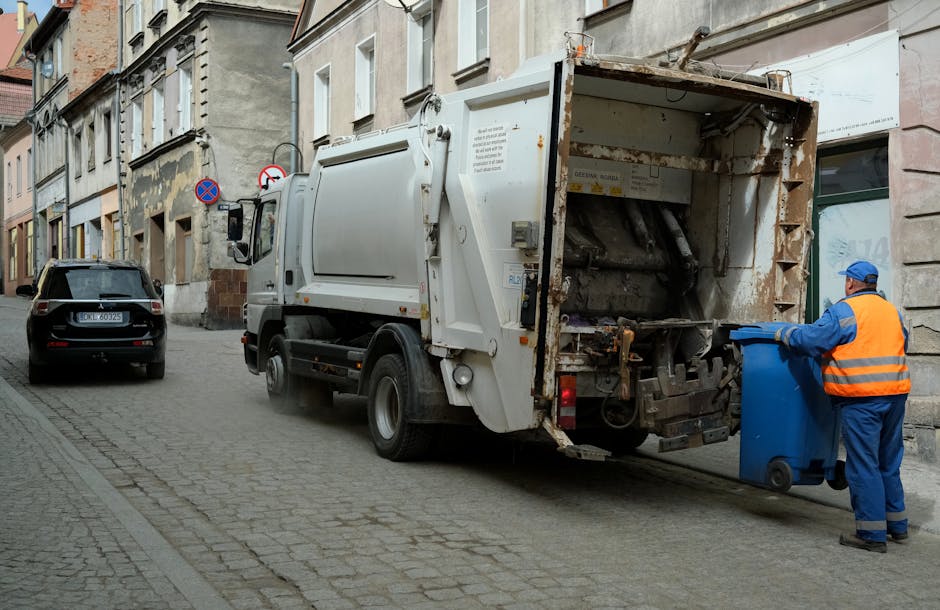 A white waste collection truck is situated on a cobbled street alongside multi-story buildings, actively engaged in rubbish removal. The rear compartment of the truck, which is open, reveals rusted, weathered metal components and a collection mechanism used for compacting or lifting waste. To the right of the truck, a worker dressed in a high-visibility orange safety vest, blue uniform, and blue cap is placing a large blue wheeled bin into the truck's holding area, indicating on-site rubbish collection. The scene includes a dark-colored car parked further down the street on the left side, and environmental elements such as street signs and a narrow urban pavement enhance the setting. Bright, natural daylight illuminates the scene, emphasizing the textures of the cobblestones, the metal surfaces, and the worker’s safety attire. The image illustrates a typical example of private rubbish collection service in an urban environment, aligning with the themes of independent waste handling and rubbish removal at residential or commercial premises.