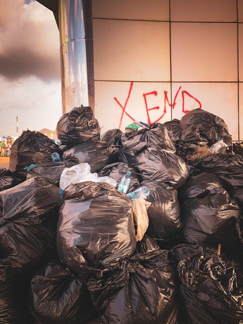 A white waste collection truck is situated on a cobbled street alongside multi-story buildings, actively engaged in rubbish removal. The rear compartment of the truck, which is open, reveals rusted, weathered metal components and a collection mechanism used for compacting or lifting waste. To the right of the truck, a worker dressed in a high-visibility orange safety vest, blue uniform, and blue cap is placing a large blue wheeled bin into the truck's holding area, indicating on-site rubbish collection. The scene includes a dark-colored car parked further down the street on the left side, and environmental elements such as street signs and a narrow urban pavement enhance the setting. Bright, natural daylight illuminates the scene, emphasizing the textures of the cobblestones, the metal surfaces, and the worker’s safety attire. The image illustrates a typical example of private rubbish collection service in an urban environment, aligning with the themes of independent waste handling and rubbish removal at residential or commercial premises.
